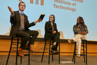 Ben Armstrong, Yvonne Hao, and Sienna Leis sit on stools in front of a large projection screen that says "Massachusetts Institute of Technology" on it. Armstrong is gesturing and speaking while the others listen.