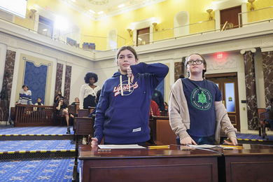 Photo of two middle school girls standing behind a desk in a replica of the U.S. Senate chambers. One girl shows her thumb pointing down for a "nay" vote.