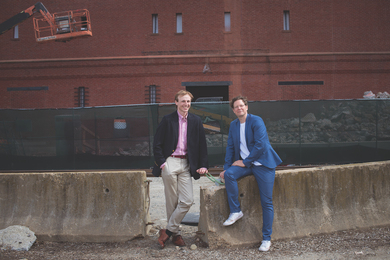 Christoph Reinhart and Zachary Berzolla lean against a concrete Jersey barrier in front of a construction site. 