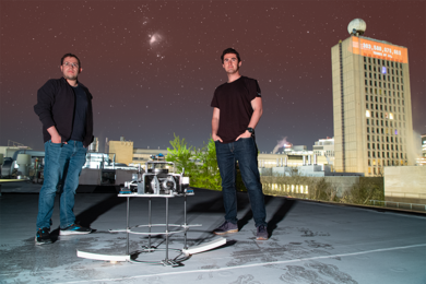 Alejandro Cabrales and Evan Kramer stand on a rooftop with the SpinAp space telescope hardware testbed between them. A purple/blue night sky can be seen in the background. To the right, a tall building is seen with an orange countdown clock illuminated toward the top with a number in the 300 billions appearing.