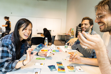 3 people smile excitedly while playing a card game. The game’s cards have blue waves on them, and there is colorful money.