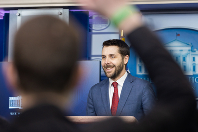 Brian Deese speaks in the White House Briefing Room. In the blurry foreground, a person raises their hand.