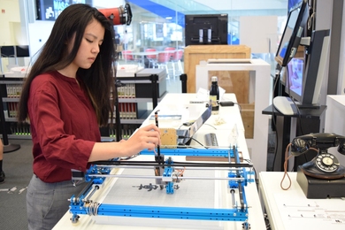 Mengying Cathy Fang stands at a workbench using a blue calligraphy plotter in a lab space