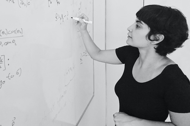 Black-and-white photo of Leila Mirzagholi standing at a whiteboard, writing equations.