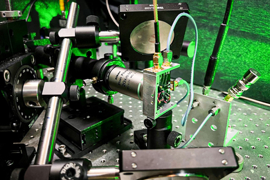 A closeup of the laser equipment shows metallic rods, a circuit board, wires, and lenses, all in a green glow.