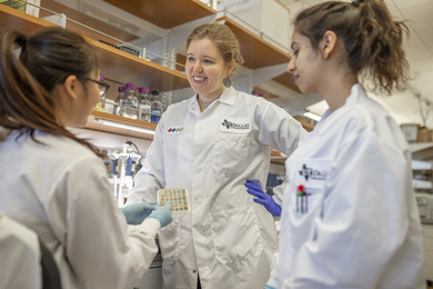 Nicole De Nisco and two others chat at a biology lab bench. All three are wearing white lab coats and hair in ponytails.