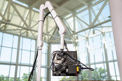 The Noncontact Laser Ultrasound optical head, a collection of circuits and wires on a black square pegboard, is suspended at the end of a robotic arm inside a large atrium filled with windows looking toward a blue sky.