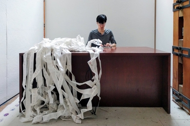Jonathan Zong is sitting at a desk, typing on a keyboard connected to a printing machine delivering large amounts of paper where letters of different sizes are visible.