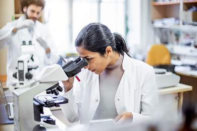 A lab worker looking through a microscope with lab and another lab worker in blurry background.
