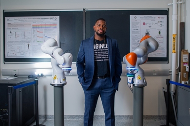 Michael West, wearing a suit with an "Engineer" T-shirt, stands in front of chalkboard, flanked by two robotic arms.