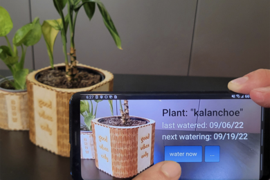 A housplant inside a wooden pot with "good vibes only" on it is seen both in the background and in a cellphone screen that is imaging the scene. On the cellphone version of the scene, overlaid text says "Plant: Kalanchoe. Last watered: 09/06/22, Next watering: 09/19/22, with a button that says "Water now" underneath 