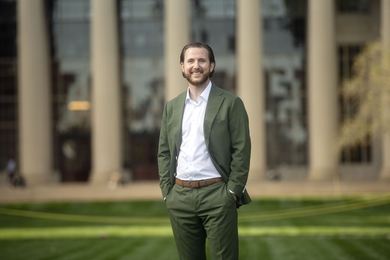 Drew Story stands on MIT's Killian Court on a sunny day, with grass and five columns of Building 10 visible behind him