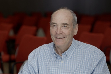 Mark Bear is sitting in an auditorium full of red chairs and smiling.