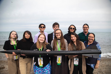 10 people standing on a beach pose with their black rocket, which they hold horizontally. It's a cloudy day.