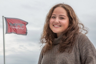 MIT Senior Tatum Wilhem is pictured close up, outdoors against a gray sky, with an MIT Crew flag flying in the background.