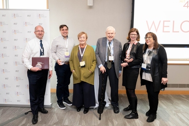 Six people wearing lanyards pose on a stage. One holds an award.