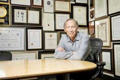 Robert Langer sits at an empty table. The walls behind him are covered with framed awards and plaques