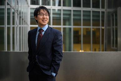William Li stands outside and looks to the right. Rows of windows are in the background.
