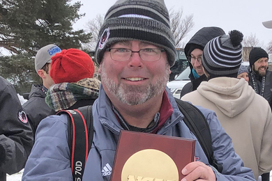 Photo of Ken Johnson outside in cold-weather gear holding a large NCAA trophy, while about others gather behind him, talking to each other