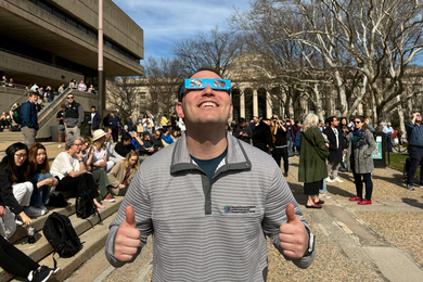 Adam Pressel wears special eclipse glasses as he looks up to the sky, with many people in background.