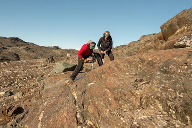 On a vast rocky landscape, Claire Nichols drills into brown rock as Ben Weiss smiles. Both wear protective goggles.