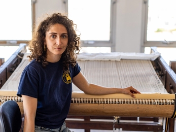 Chloe Bensahel sits in front of a loom, looking at the camera