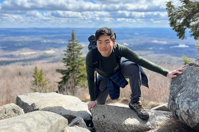 A young man wearing a long-sleeve T-shirt, jeans, and sneakers scrambles over a rocky ledge atop a high mountain. Clouds, a broad sky, and forested hilltops are visible in the background.