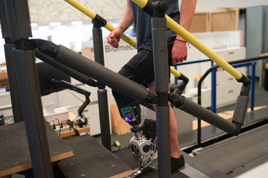 A person wears a prosthetic leg with a circuit board while walking up stairs in a lab.