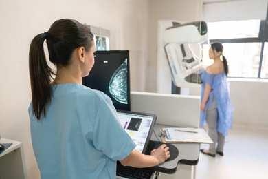 A doctor looks at breast Xray with patient and scanner in background.