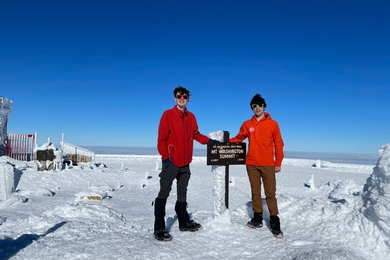 Nathanael Jenkins and Timur Uyumaz pose with the summit marker on Mt. Washington on a sunny day. They wear cold-weather gear and there is snow on the ground.