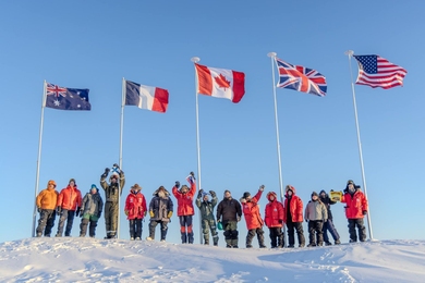 On a sunny day in the Arctic, 15 people bundled in cold-weather gear stand in a line with flags for Australia, France, Canada, United Kingdom, and USA waving on poles above them.