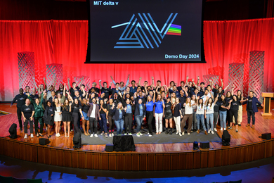 About 70 people raise their hands in celebration on a stage. A banner above them reads “MIT delta v Demo Day 2024.”