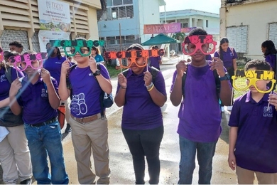 6 students wearing matching uniforms hold colorful glasses labeled with STEAM subjects, "Science," "Technology," "Engineering," "Arts," and "Math," in front of a school building
