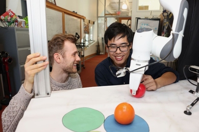 Vincent Sitzmann and Boyuan Chen place a toy apple in front of a robotic arm attached to a table. A toy orange is nearby, placed on top of a blue circular mat, near light and dark green mats.