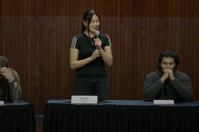 Claire Wang stands on stage with a microphone and two other competitors are seated.