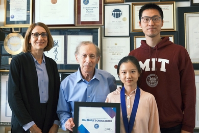  Ana Jaklenec, Robert Langer, Linzixuan (Rhoda) Zhang, and Xin Yang stand in front of a wall of framed awards. Zhang wears a medal ribbon around her neck and, together with Langer, holds a framed certificate. 