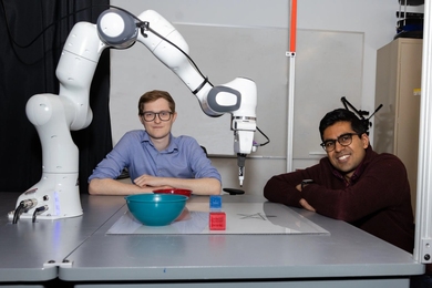 Two students sit at a table dominated by a white robotic arm that drew a star on a whiteboard.