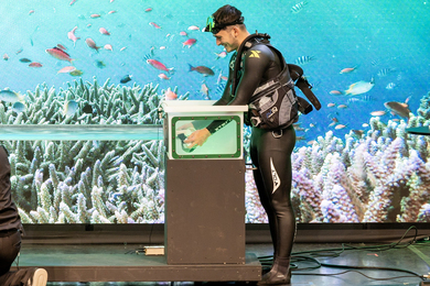 A scuba diver puts goggles in an aquarium while on stage