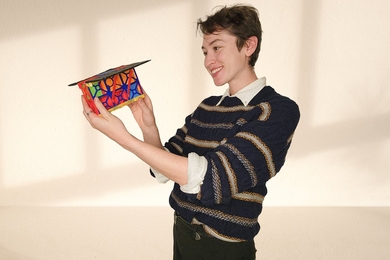 Sydney Dolan holds a 3D-printed satellite model, a bit smaller than a shoebox, in front of a white wall