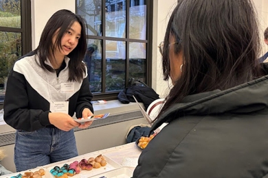 Angelina Wu, standing at a table laden with pastries, prepares to hand a brochure to someone