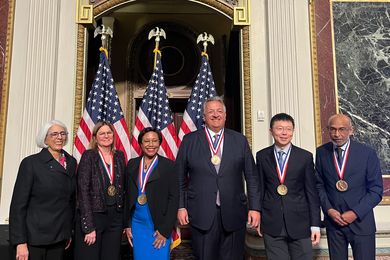 Arati Prabhakar, Angela Belcher, Paula Hammond, Noubar Afeyan, Feng Zhang, and Emory Brown pose together in front of 3 U.S. flags..