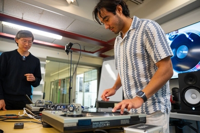 Philip Tan guides student Karl Velazquez, who is standing over a turntable with his finger on the platter