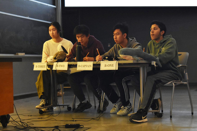 Katherine Wang, Evan Zhang, Yunyi Ling, and Kian Dhawan sit at a table holding pencils in a lecture hall. There are plush animals and a water bottle on the table. The students are focused on something left of the field of view.