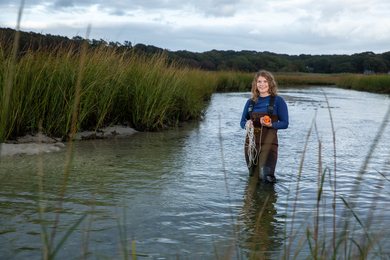 Faith Brooks, wearing waders stands knee-deep in a pond, holding an instrument and a coil of rope