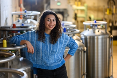 Zoe Fisher stands in a lab space in front of several large stainless steel dewers