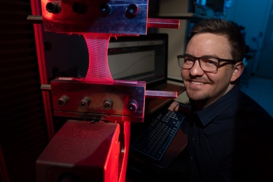 Steve Gillmer poses with an industrial knitting machine in a darkened room with red and blue light.
