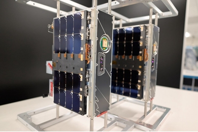 On a tabletop, two small boxy satellites, roughly 20 x 10 x 30 cm in size. 