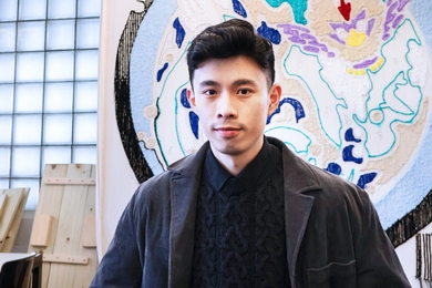 A young man with short black hair wearing a dark cable-knit sweater, black collared shirt, and corduroy jacket stands in front of a large abstract textile artwork featuring bold shapes in blue, white, and purple.