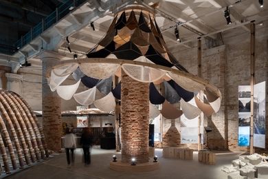 Architectural installation with layered panels of fabric, wood, and bio-based materials suspended from a brick column resembles a giant lampshade. 