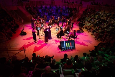 Top-town view of 18 musicians playing on a circular stage under red light, surrounded by an audience